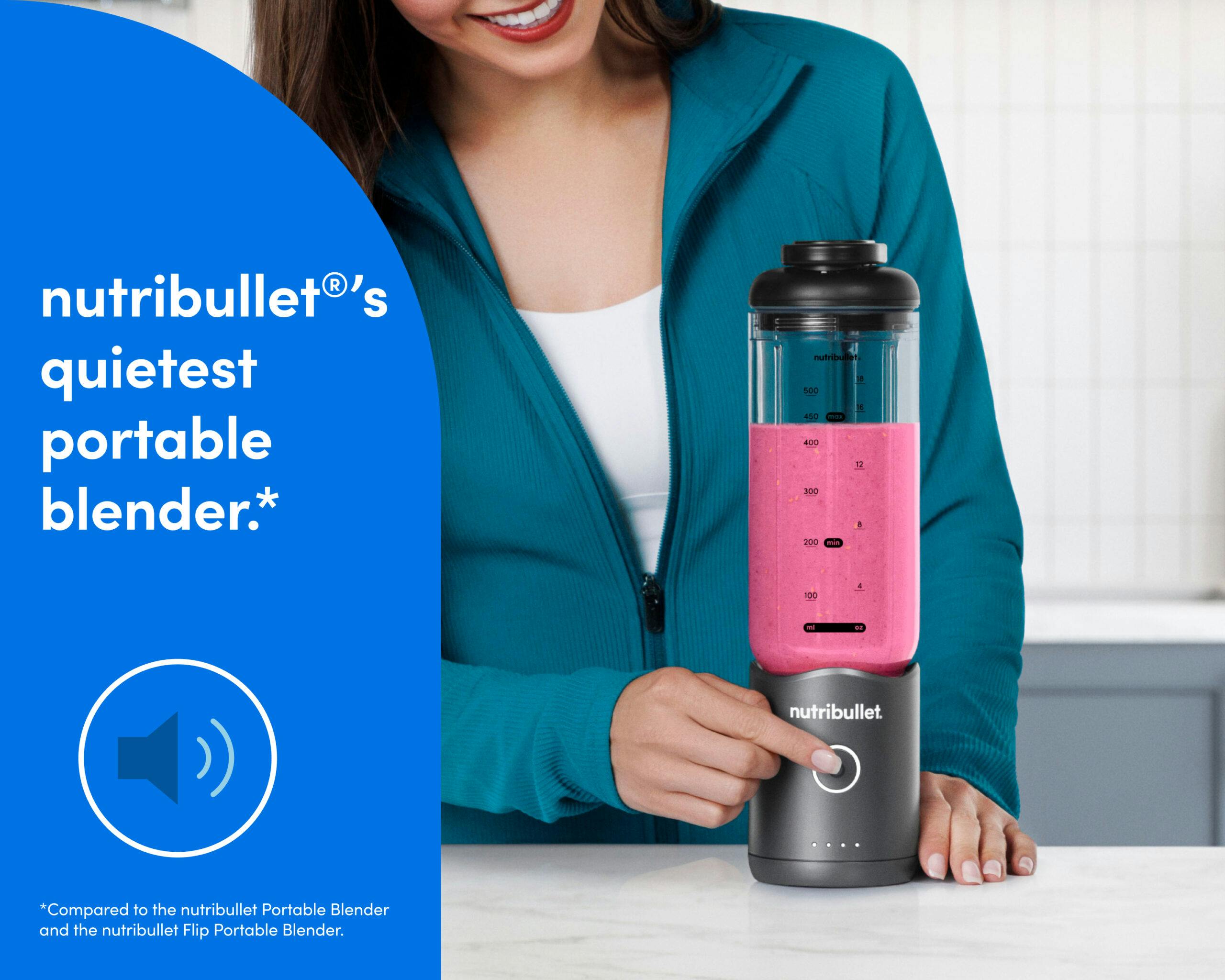 A woman in a teal zip-up athletic jacket presses the blend button on the bottom of the motor base to blend a bright pink smoothie.