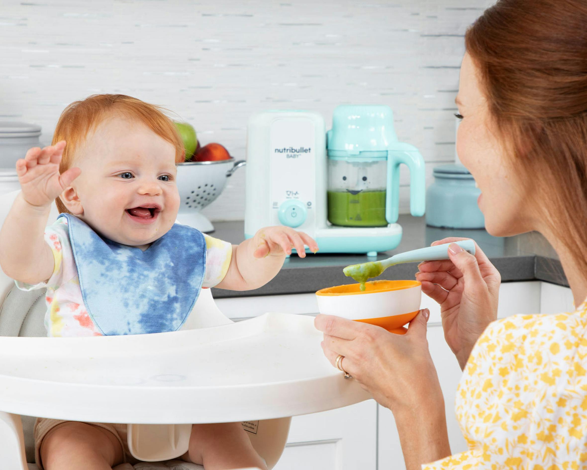 Mom feeding baby in a high chair with the baby steam and blend in the background