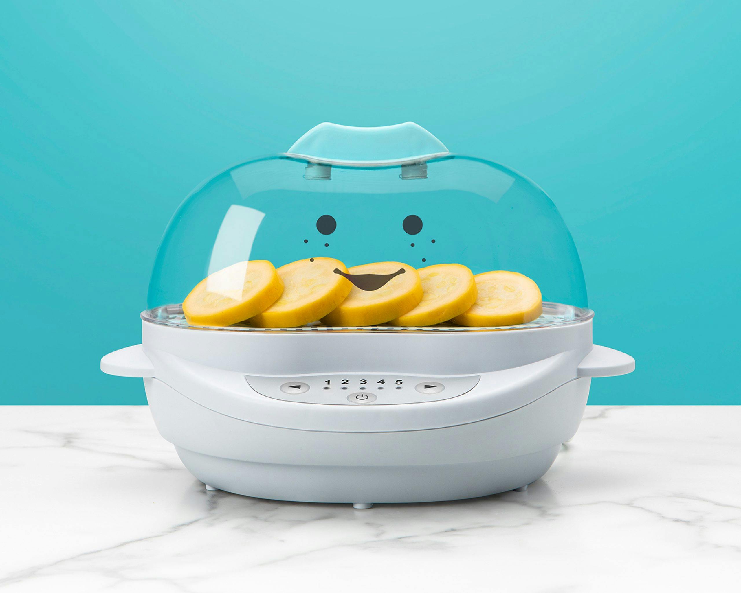 White NutriBullet Baby Steamer filled with sliced veggies with teal accents with smiley face on teal background.